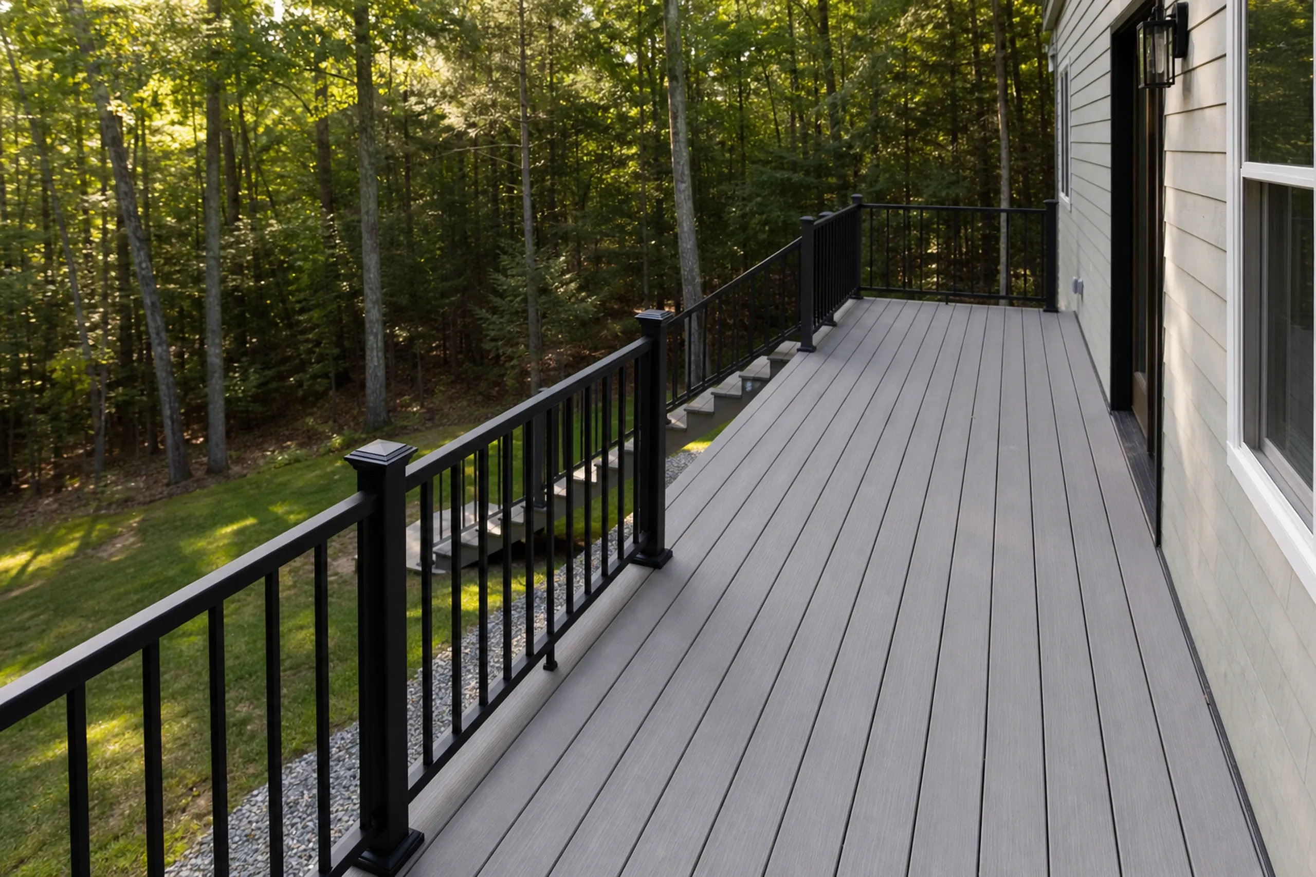 Empty gray wooden deck with black metal railing attached to gray house exterior in a wooded area with stone steps below.