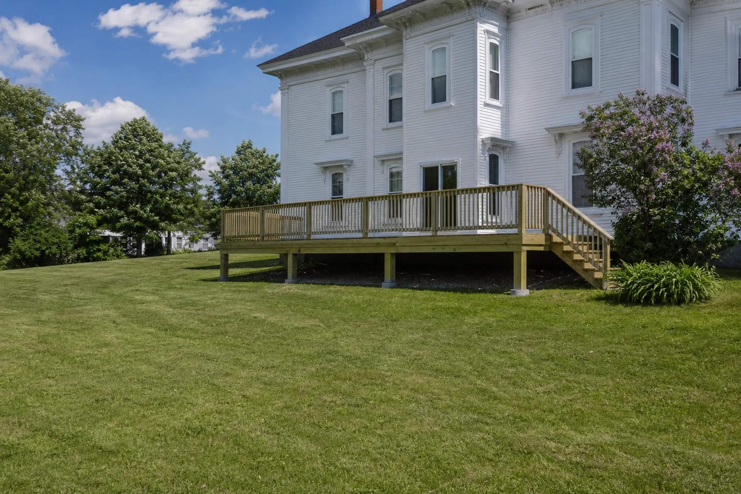 A white two-story house with a wooden deck and railing on a grassy lawn, with no people visible.
