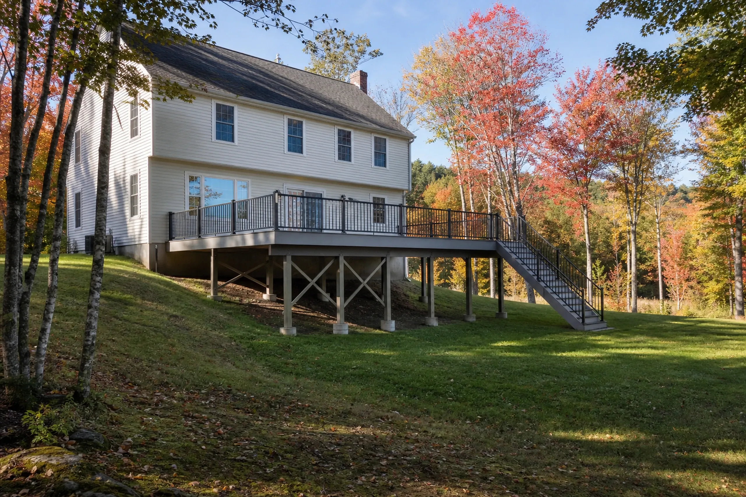 White two-story house with an elevated deck featuring black metal railing and stairs, surrounded by autumn trees and grass.