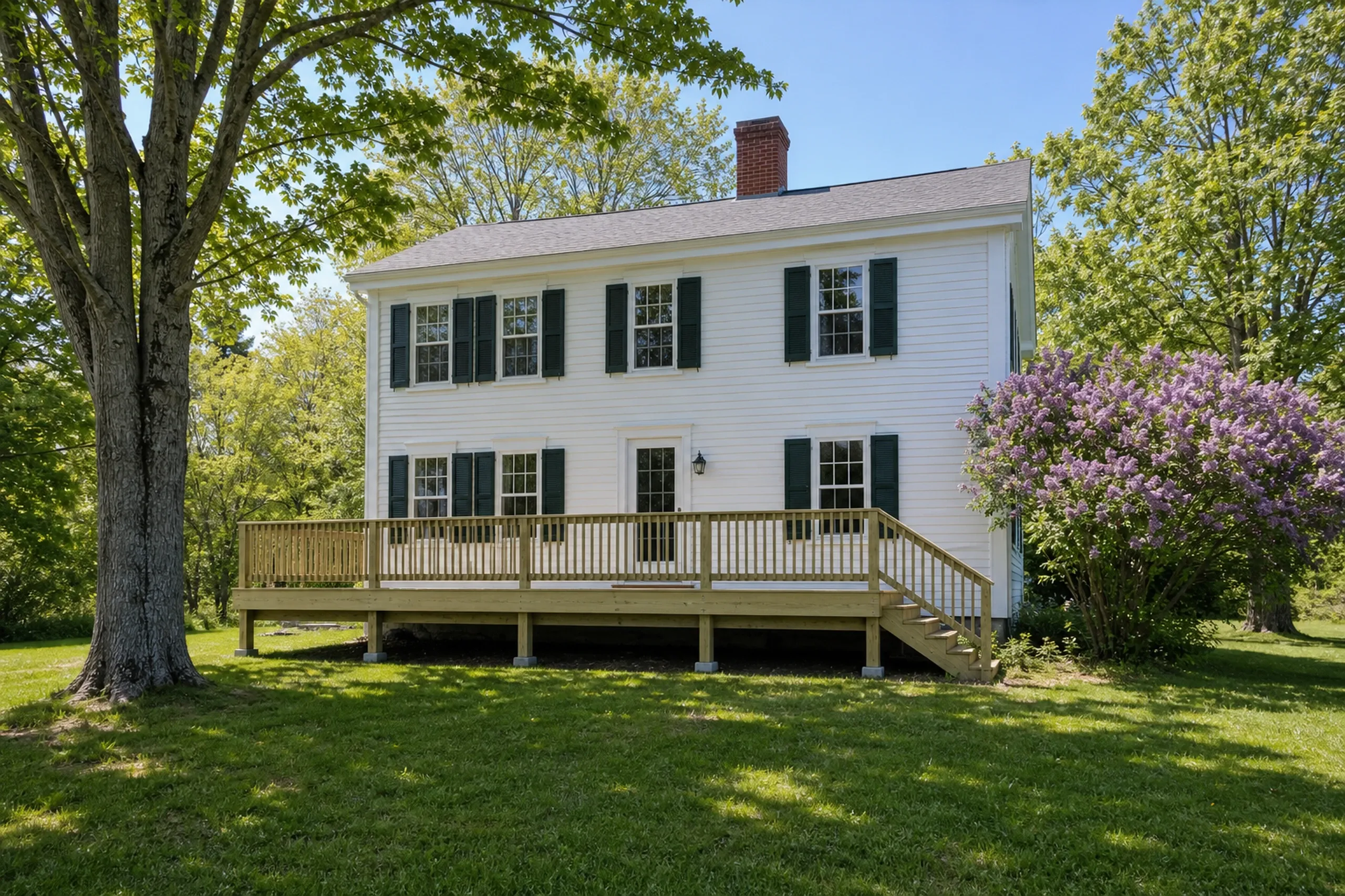 White two-story house with green shutters, wood deck and railing, surrounded by grass, trees, and a purple-flowered bush; no people visible.