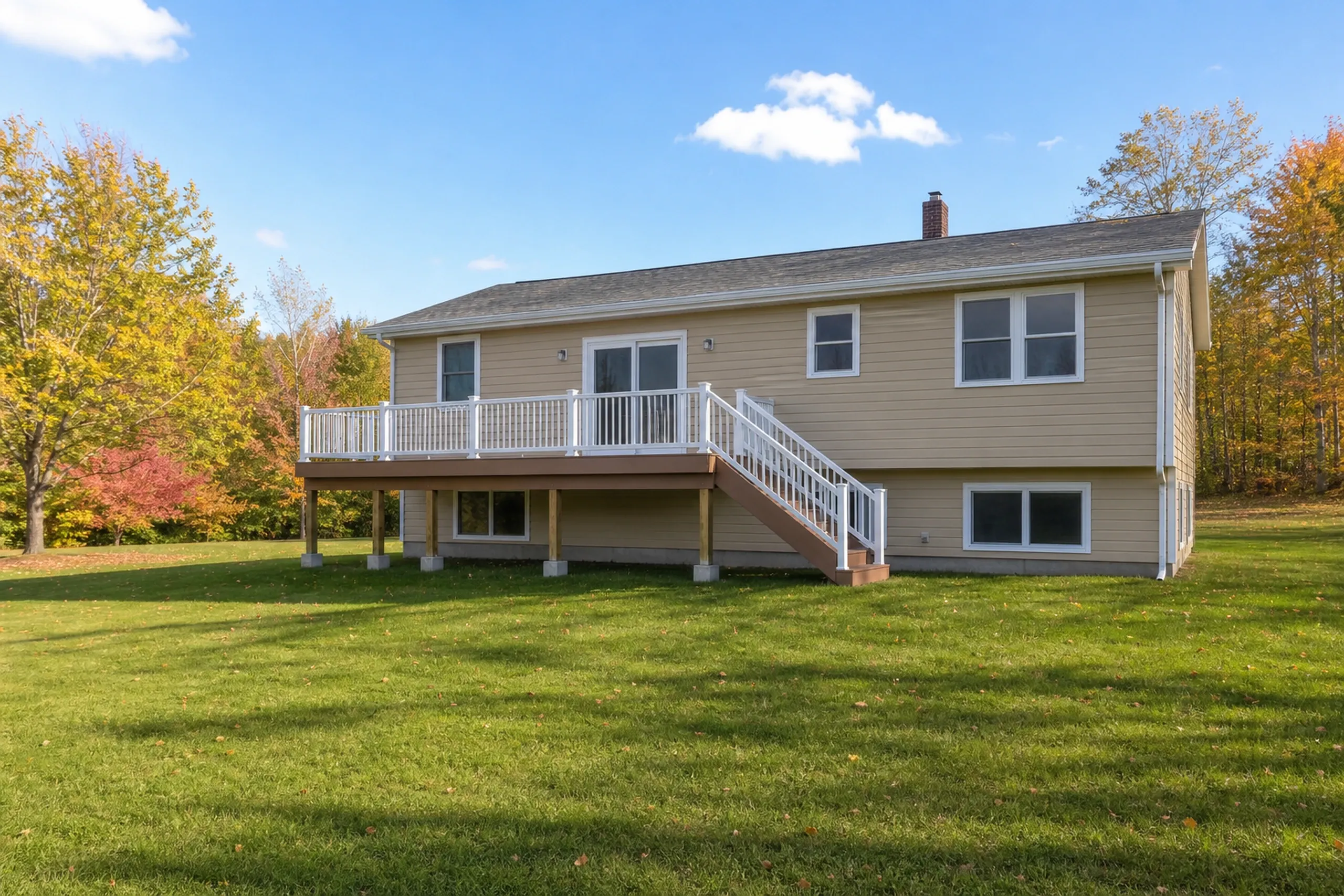 Beige single-story house with white railing deck and stairs, surrounded by green lawn and trees with autumn foliage.
