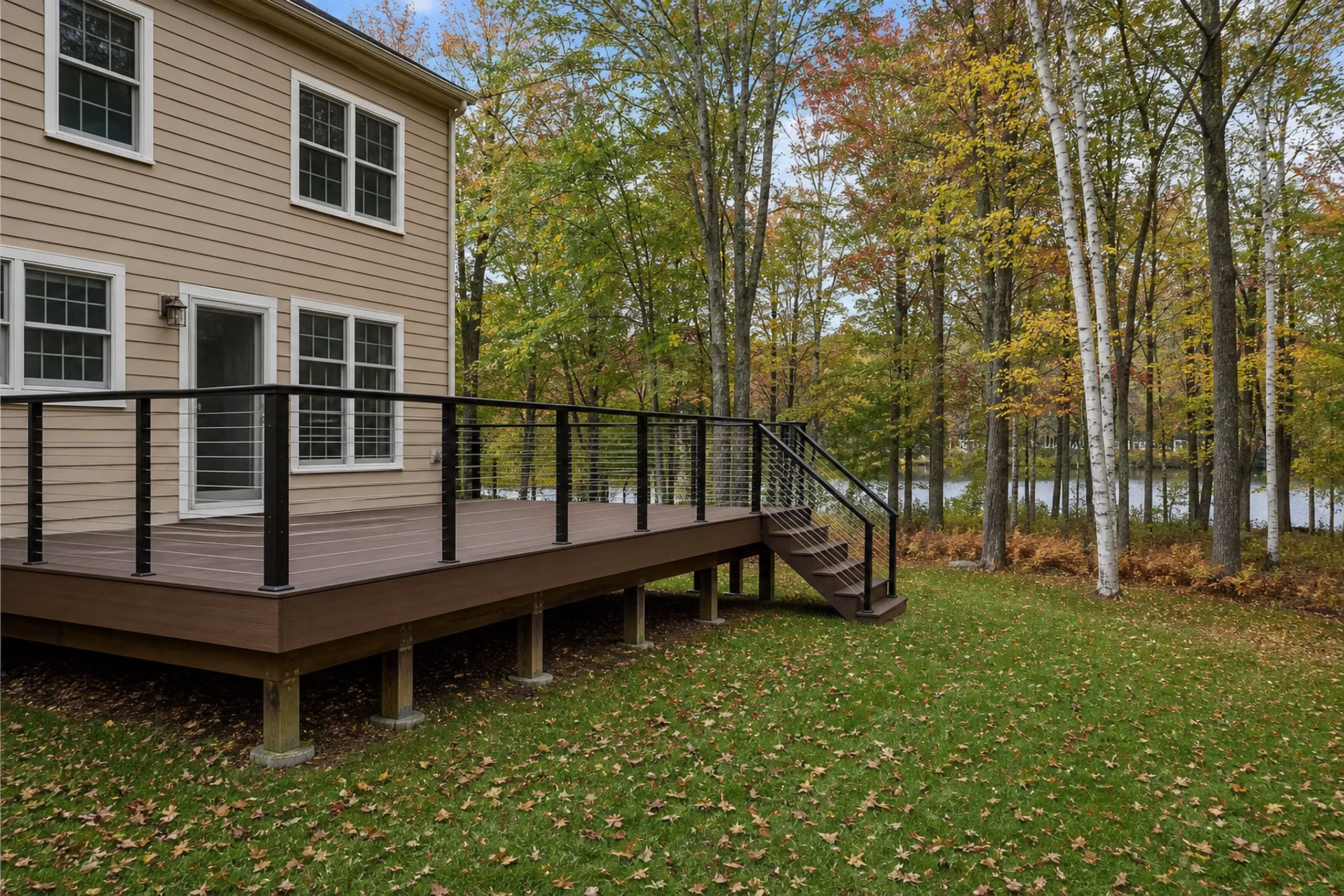 A beige two-story house with a raised brown deck featuring black metal railings and stairs, overlooking a tree-lined grassy yard.